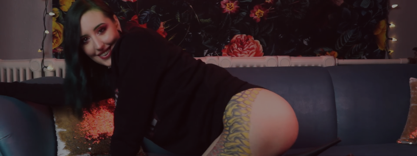 Tattooed Pale Alternative Goth Punk Domme Miss Faith Rae kneels sideways on her turquoise couch, her head turned to the side to look at you through the camera. She wears tall black high heels, and a black sweater. Her perfect ass sticking out and showing off the reptile snakeskin animal print tattoo that frames her perfect peach ass and goes down the side of her thigh. Daddy Faith Rae has green hair with matching eyebrows. Her eye makeup is red and gold, and her lips are a deep berry pink. You want to give Miss Faith Rae everything.